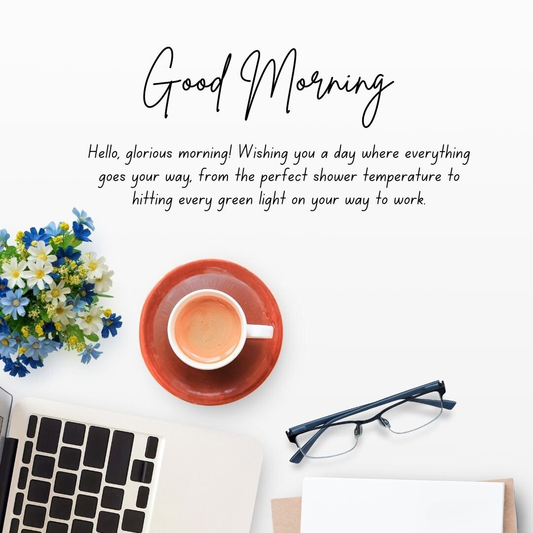 Funny Good Morning Wishes on a desk setup featuring a red coffee cup, laptop, glasses, and a bouquet of fresh blue and white flowers, complemented by a humorous morning wish for hitting every green light on the way to work.