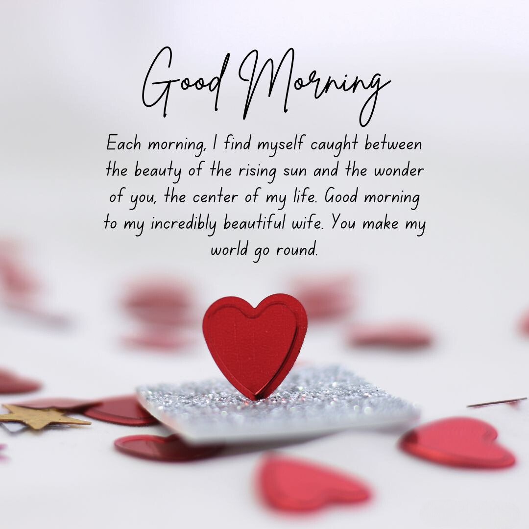 A red heart decoration stands on a glittery surface surrounded by small red heart-shaped confetti. The image includes the text 'Good Morning' and 'Each morning, I find myself caught between the beauty of the rising sun and the wonder of you, the center of my life. Good morning to my incredibly beautiful wife. You make my world go round,' making it perfect for good morning messages for wife image.