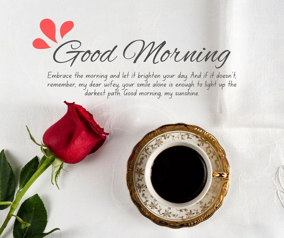 A single red rose and a cup of coffee in an ornate teacup placed on a white tablecloth. The image includes the text 'Good Morning' and 'Embrace the morning and let it brighten your day. And if it doesn't, remember, my dear wifey, your smile alone is enough to light up the darkest path. Good morning, my sunshine,' making it perfect for good morning messages for wife image.