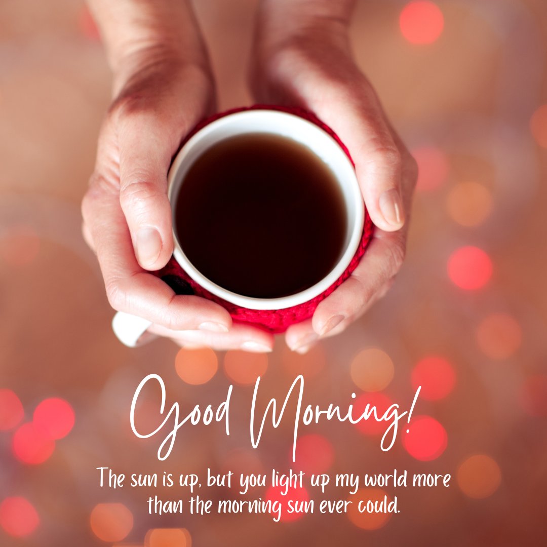 A close-up view of a person's hands holding a red mug of coffee with a warm, blurred background of glowing lights. The image also includes the text 'Good Morning! The sun is up, but you light up my world more than the morning sun ever could,' making it a perfect visual for romantic good morning messages for a wife.